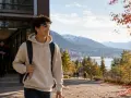 Ein junger Mann mit Rucksack verlässt ein Gebäude, im Hintergrund Berge und Stadt. Perfekt für sein Auslandsjahr Kanada.