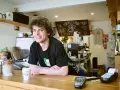 Ein junger Mann mit lockigen Haaren lächelt hinter der Theke eines Cafés. Er scheint seine **zwei Werkstudentenjobs** zu genießen, während er Kaffee zubereitet.