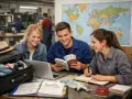Drei junge Leute planen ihre berufsausbildung usa vergleich. Sie sitzen an einem Tisch mit einer Weltkarte, Pässen und einem Koffer.