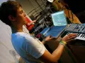 Junger Mann am Laptop und Keyboard, der Musik produziert. Ein Schlagzeuger im Hintergrund. Perfekt für ein Musikproduktion Studium.