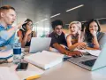 Fünf Studierende lernen gemeinsam. Sie lachen und diskutieren, während sie an Laptops arbeiten. Ein Mann gestikuliert lebhaft, was auf ein spannendes Studium im Ausland hindeutet.