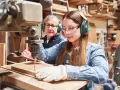 Junge Frau lernt an einer Bohrmaschine, während ein älterer Mann zusieht. Eine gute Ausbildung, welche Ausbildungen dauern 2 Jahre, ist hier vielleicht der Anfang.