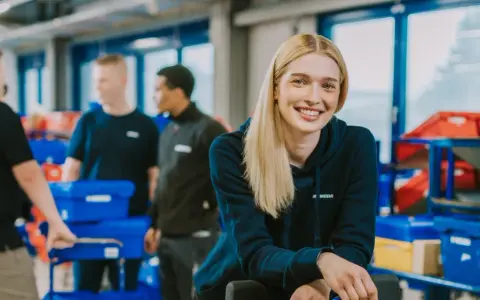 Lächelnde junge Frau im Lager, die ihren Werkstudentenjob genießt. Im Hintergrund arbeiten Kollegen mit blauen Behältern.