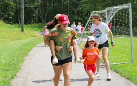Mädchen haben Spaß bei einem Sommercamp in den USA. Eine trägt die andere auf dem Rücken, während sie lachend über einen Weg laufen.