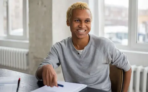 Lächelnder Mann mit blonden Haaren sitzt am Schreibtisch, Notizbuch und Stift bereit für sein Praktikum in Deutschland.