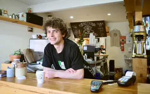 Ein junger Mann mit lockigen Haaren lächelt hinter der Theke eines Cafés. Er scheint seine **zwei Werkstudentenjobs** zu genießen, während er Kaffee zubereitet.