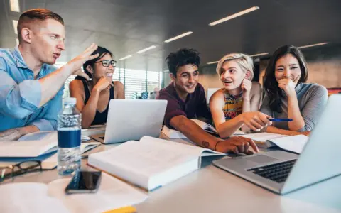 Fünf Studierende lernen gemeinsam. Sie lachen und diskutieren, während sie an Laptops arbeiten. Ein Mann gestikuliert lebhaft, was auf ein spannendes Studium im Ausland hindeutet.