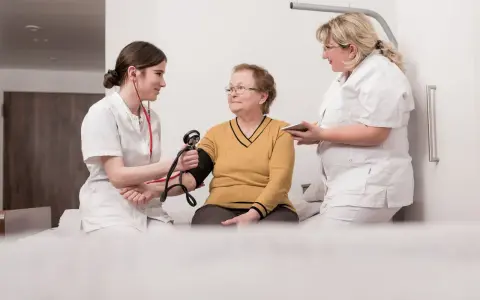Zwei Pflegekräfte messen einer älteren Frau den Blutdruck. Ein Einblick in die Praxis des Rehabilitationspsychologie Studiums.