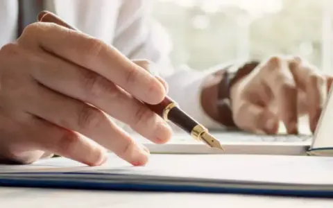 Person schreibt mit Füllfederhalter auf Dokumenten, während er am Laptop arbeitet. Ein gutes Beispiel für einen Praktikumsbericht.