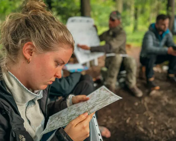 Frau studiert Karte, während Männer im Hintergrund bei einer Ausbildung im Wald sitzen. Solche intensiven, kurzen **ausbildungen die nur 1 jahr dauern** sind ideal für schnelle Karriereschritte.