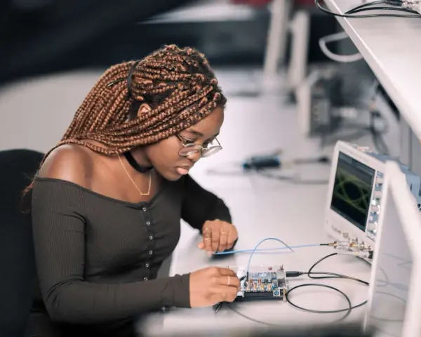 Junge Frau mit geflochtenen Haaren arbeitet konzentriert an einer Platine, während ein Oszilloskop im Hintergrund läuft. Ein spannendes elektrotechnik studium.