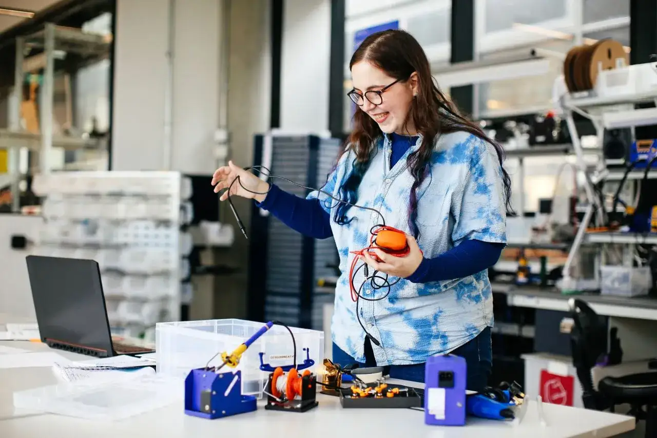 Junge Frau mit Brille lacht und hält ein orangefarbenes Gerät mit Kabeln. Sie arbeitet in einem Labor, ein Beispiel für kreative Berufe.