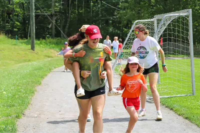 Mädchen haben Spaß bei einem Sommercamp in den USA. Eine trägt die andere auf dem Rücken, während sie lachend über einen Weg laufen.