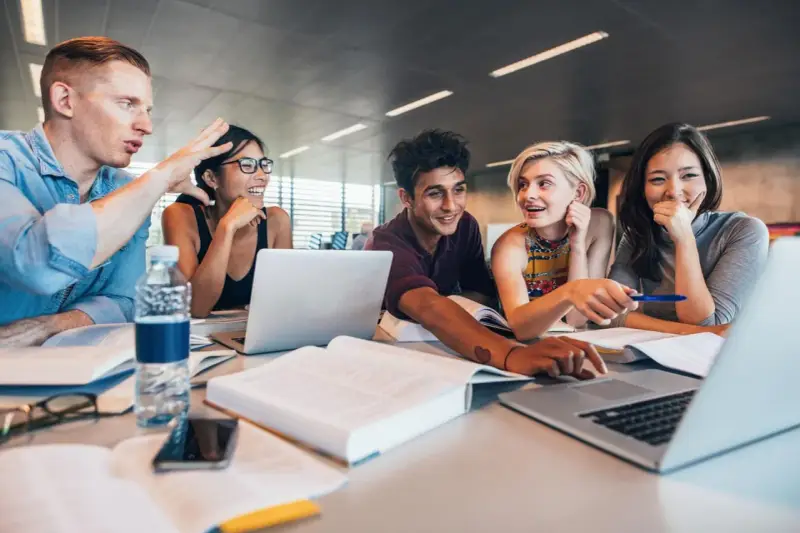 Fünf Studierende lernen gemeinsam. Sie lachen und diskutieren, während sie an Laptops arbeiten. Ein Mann gestikuliert lebhaft, was auf ein spannendes Studium im Ausland hindeutet.