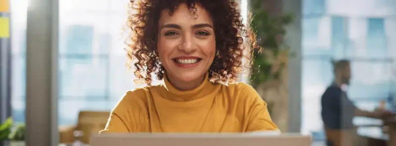 Lächelnde Frau mit Locken am Laptop, bereit für ihre Bewerbung als studentische Hilfskraft.